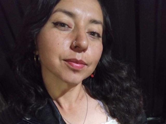 DouglasAndVioleth with wavy black hair, nose ring, black eyeliner, pink lips, small red heart earring, and black leather jacket, smiling slightly. No visible tits or ass.