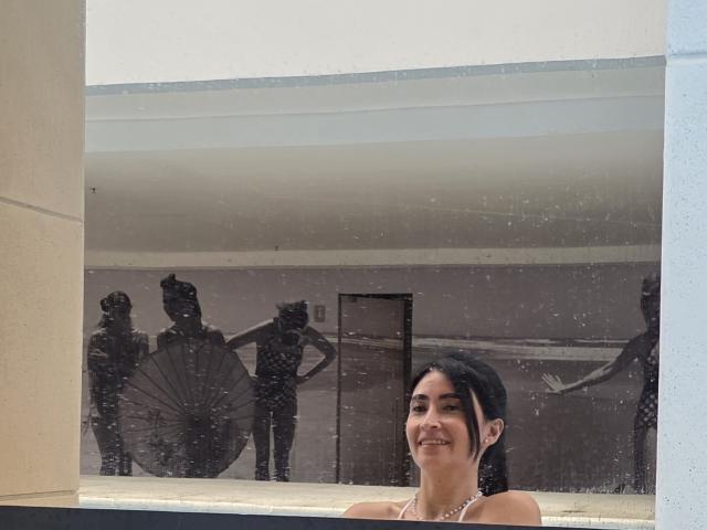 JainaRogers with dark hair, smiling, has a small nose, wearing a necklace, reflected in a dirty glass window with black silhouetted figures in the background.