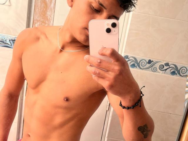 JamessHott sin camisa, con un cuerpo delgado y tonificado, pequeños pezones marrones y un tatuaje en su antebrazo derecho, tomando una selfie con un iPhone blanco..