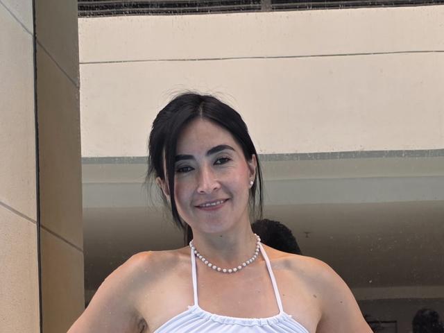 JainaRogers has dark hair, a small smile, and is wearing a white halter top with a pearl necklace, showing her bare shoulders and a small tattoo on her right arm.