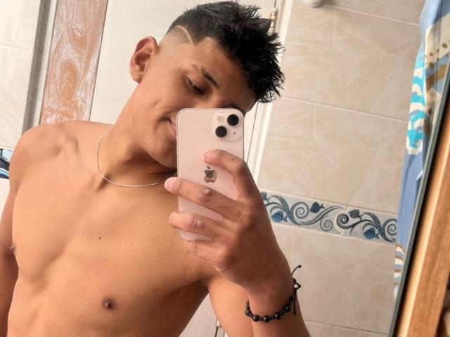 Shirtless JamessHott with a cute, slim build, short black hair, and a silver necklace, licking an iPhone in a bathroom mirror selfie. His cock is not visible.
