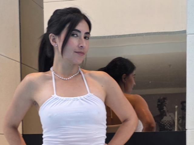 JainaRogers with dark hair in a ponytail, wearing a white halter top and pearl necklace, standing in front of a mirror, showing her small tits and subtle smile.