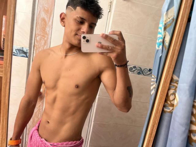 Shirtless JamessHott with a slim, toned physique, short black hair, and a slight smirk, taking a mirror selfie, wearing pink pants, wearing an orange bracelet, and a necklace.