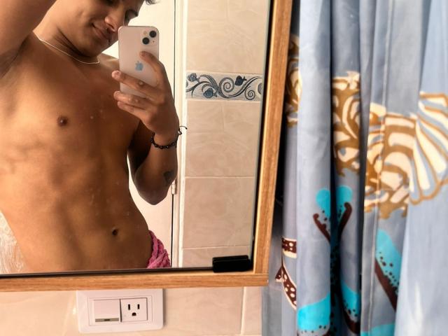 A mirror selfie of a shirtless JamessHott with a slim, toned physique. He has light brown skin, a slight smile, and short, dark hair. He holds a white phone with a blue circular button in his right hand. He wears pink shorts. The reflection shows his defined abs and a black bracelet on his left wrist. The mirror has a wooden frame, and a blue shower curtain with brown and white patterns is to the right. A light switch is at the bottom.