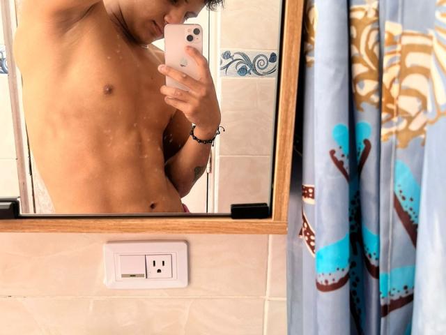 Selfie no espelho de JamessHott sem camisa, com um torso tonificado e musculoso, segurando um celular, mostrando abdominais definidos e um leve bronzeado. Cortina de chuveiro azul e marrom à direita..