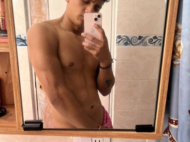 Mirror selfie of JamessHott, a young, slim, shirtless man with light brown skin and a small, flat chest. He holds a pink iPhone with a white case in his right hand, slightly tilting his head. He wears pink, patterned underwear and has a tattoo on his left forearm. The mirror has a wooden frame and a black, rectangular mount. The reflection shows his toned, muscular abdomen. His curly, dark brown hair is slightly messy. His left arm has a black bracelet. The background is a bathroom with beige tiles and a decorative blue pattern.