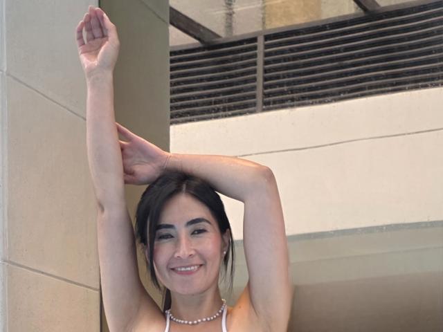 JainaRogers has her arms raised, showing off her bare tits, and is smiling with dark hair and a pearl necklace. She's leaning against a beige wall.