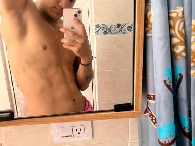 Mirror selfie of a shirtless JamessHott with a slim, toned, tan body, small nipples, and a visible tattoo on the left wrist, wearing pink underwear.