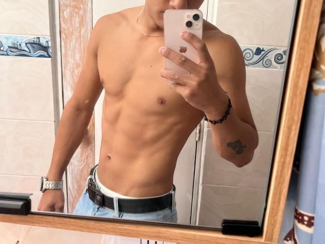 JamessHott sem camisa, com abdominais definidos, vestindo jeans azuis e um cinto preto, tirando uma selfie no espelho com um iPhone rosa. Ele tem uma tatuagem no antebraço direito..