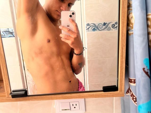 O espelho reflete o corpo superior musculoso e sem camisa de James Hott, mostrando um abdômen plano e musculoso, pequenos mamilos e uma tanga rosa com o bumbum à mostra. Ele está segurando um telefone branco..