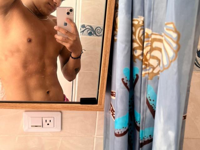 Mirror selfie of shirtless JamessHott with toned abs, holding phone, showing chest and lower face, standing in bathroom with blue patterned curtain, beige tiles, and white electrical outlet.