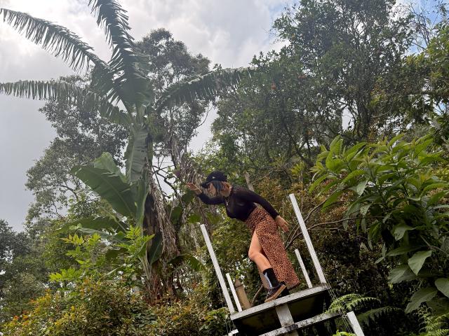 SaraleePoison with brown hair, black top, and orange patterned pants, standing on a white structure, reaching up to tall banana leaves, surrounded by lush green jungle.