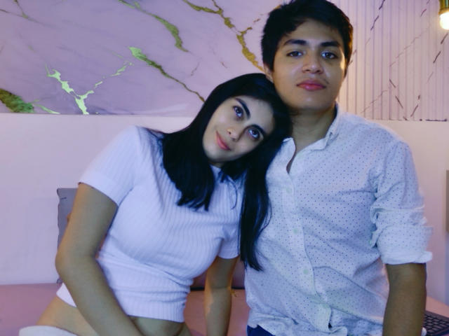 SofiNdLogan with dark hair and white top leans on a guy with short black hair and polka-dot shirt, both with serious expressions, in a dimly lit room.