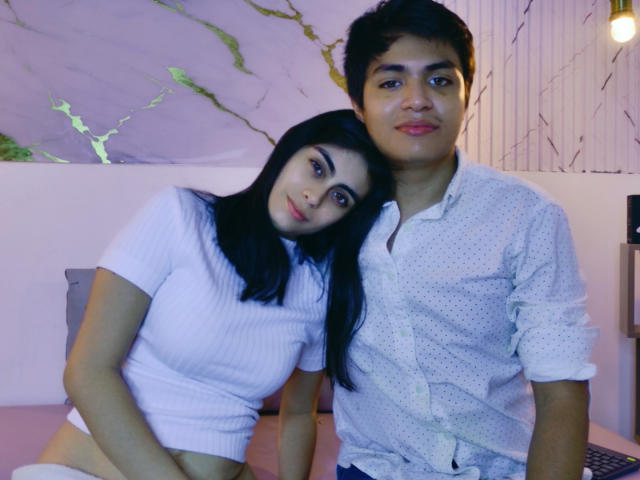 SofiNdLogan with long black hair and small tits leans on a guy with dark hair in a white polka dot shirt, both smiling in a pink-lit room.