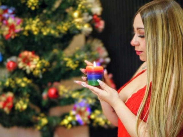 ImXNicole, avec de longs cheveux blonds, une robe rouge, tenant une bougie colorée, debout près d'un sapin de Noël décoré, se concentrant sur la bougie, montrant légèrement ses seins..