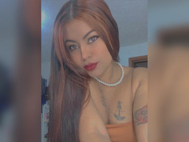 DannaHarmonyy with long brown hair, red lipstick, pearl necklace, exposed orange strap top, tattoo on chest, tattoo on arm, looking at camera with seductive gaze, bedroom setting.