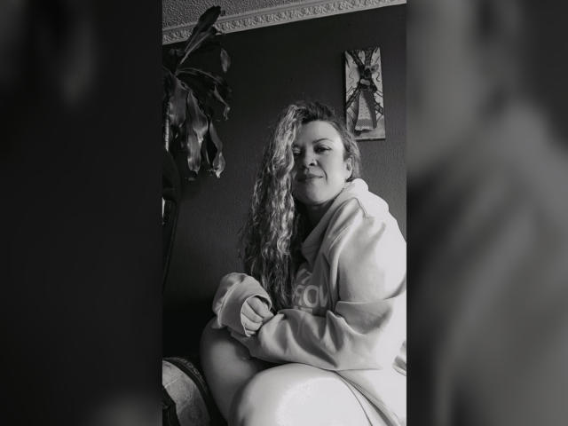 Foto in bianco e nero di RosseHot69 con i capelli ricci, che sorride, indossa una felpa larga, seduta con le ginocchia sollevate, mani sulle ginocchia, figura sfocata in primo piano, pianta e decorazioni murali sullo sfondo..