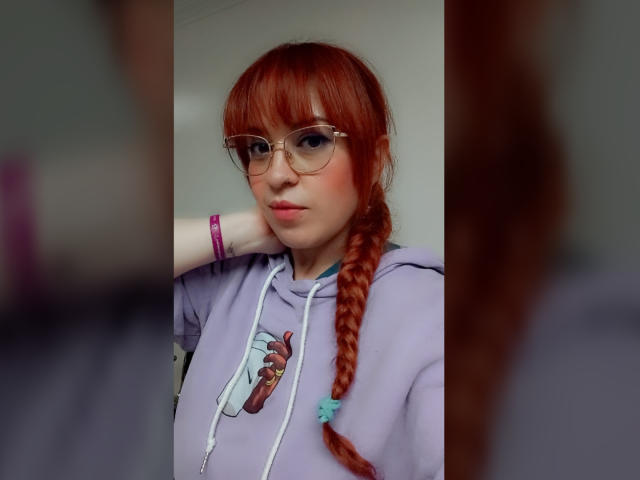 RosseHot69 has red hair in a braided ponytail, wearing clear glasses, and a light purple hoodie with a cartoon character on it. She's leaning against a wall with her right arm, wearing purple and pink bracelets. Her expression is neutral, with slight pink blush on her cheeks. The image is a selfie, with a blurred reflection on the right. The focus is on RosseHot69's face and upper body. The colors are soft, with a contrast between her hair and the hoodie.
