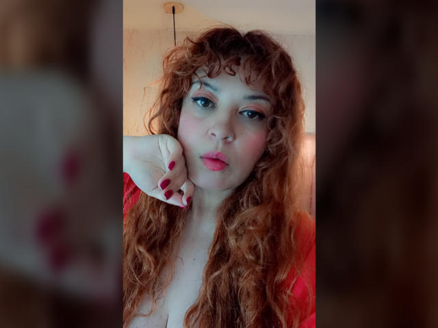 RosseHot6g has long, curly red hair and is making a pouty face. She has blue eyes, pink blush on her cheeks, and red lipstick. Her right hand with red-painted nails is resting on her cheek. She is wearing a red top. The image is a close-up, centered on her face and upper chest. The background is blurred, focusing attention on RosseHot6g. The overall color palette includes red, pink, and her natural hair color. The photograph has a soft, warm light.