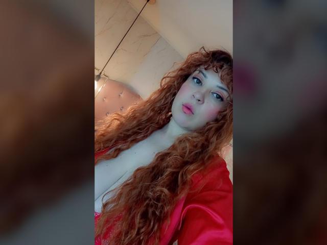 RosseHot69 con lunghi capelli rossi ricci, indossa un vestito di seta rosso, mostra il seno, ha un leggero rossore sulle guance e labbra rosa, e sta scattando un selfie..