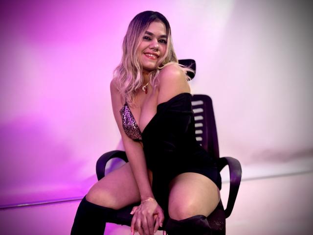 TarahDustin with blonde hair, smiling, wearing a sparkly bra, black off-shoulder top, and black knee socks, sitting on a chair with purple lighting. Boobs, ass, pussy visible.