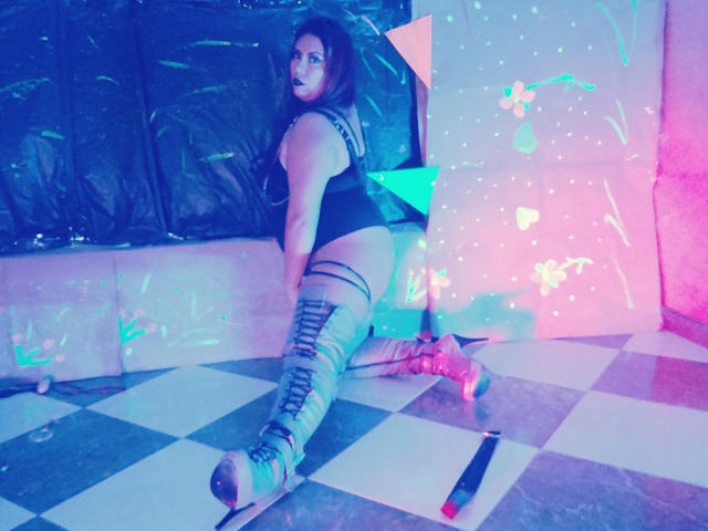 LexyDom on knees, back to camera, wearing black top, gray leggings, and roller skates, with visible ass and curly dark hair, in a checkered floor, neon-lit room.