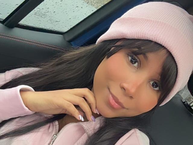 DariaEbony, avec de longs cheveux noirs, un bonnet rose et une veste zippée rose, sourit dans une voiture, se tenant le menton, avec des lèvres roses et des ongles violets. Ses seins ne sont pas visibles..