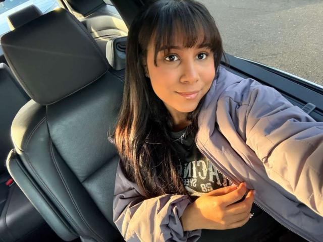Daria, com pele morena, franja preta e cabelo longo e liso, vestindo uma jaqueta roxa, sentada em um carro, sorrindo, segurando a jaqueta, com os seios levemente visíveis sob a jaqueta..