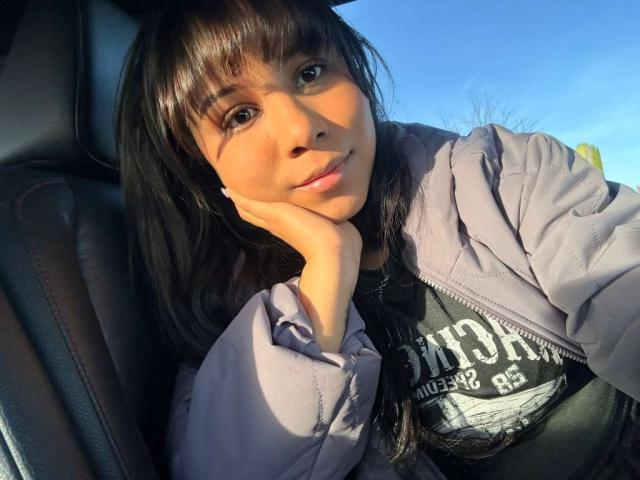 DariaEbony, con cabello oscuro y flequillo, vestida con una chaqueta morada, apoyada en su mano dentro de un coche, cielo azul afuera, luz del sol en su rostro, mirando a la cámara..