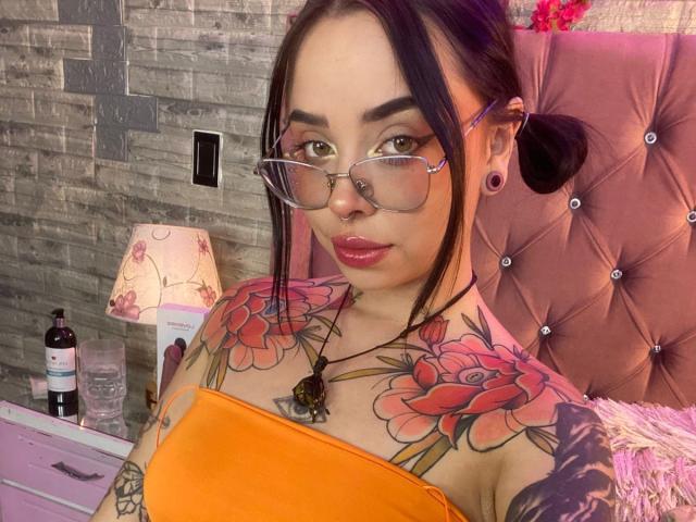 NaiaWhisper with tattoos, glasses, nose ring, and ear gauges, wearing an orange top, showing large tits, and a necklace, in a pink, tufted, headboard bedroom.