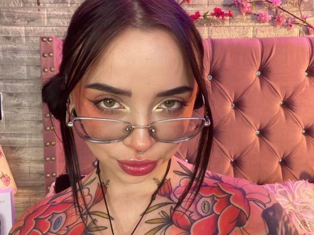 NaiaWhisper with glasses, nose ring, and red lipstick, has tattoos on her chest, and brown hair in a ponytail, looking at the camera with green eyes, and glossy pink lips.