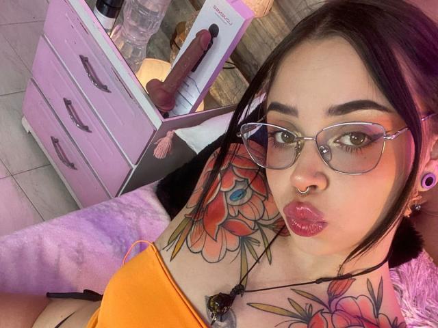 NaiaWhisper with glasses, nose ring, and pink lipstick, showing off colorful tattoos on her arm and chest, wearing an orange top, and a black necklace, with a dildo in background.