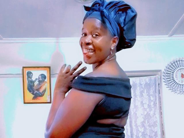 SexyBrexy with dark skin, smiling, wearing a black off-shoulder top and a large black headwrap, showing her back and hand, with visible earrings.