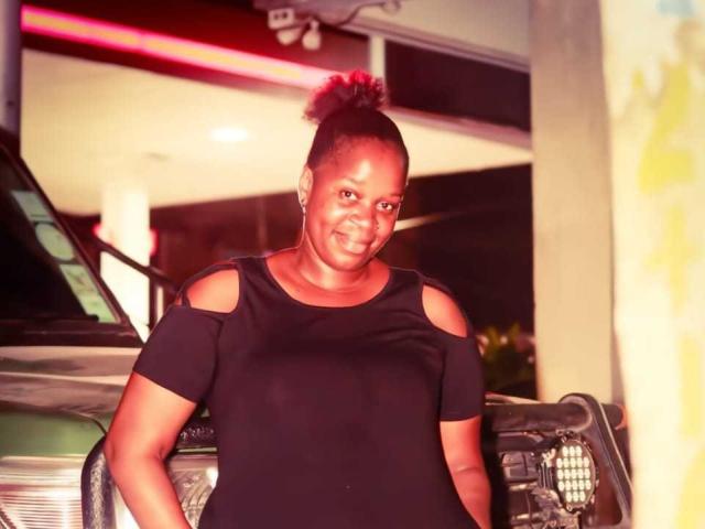 SexyBrexy with dark skin, medium build, black off-shoulder top, and short curly hair, smiling, standing in front of a green car at night.