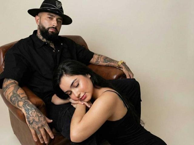 ConnieAndDarkko, bearded dude with tattoos, black hat, black shirt, sitting in brown chair, arms resting. ConnieAndDarkko, black-haired, sleeping, head on arms, black dress, relaxed.