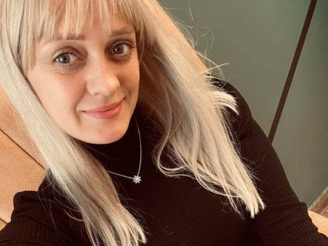 KiraHarra with blonde hair, black ribbed shirt, nose ring, and star necklace, smiling at the camera, showing her cute face and subtle pink lips.