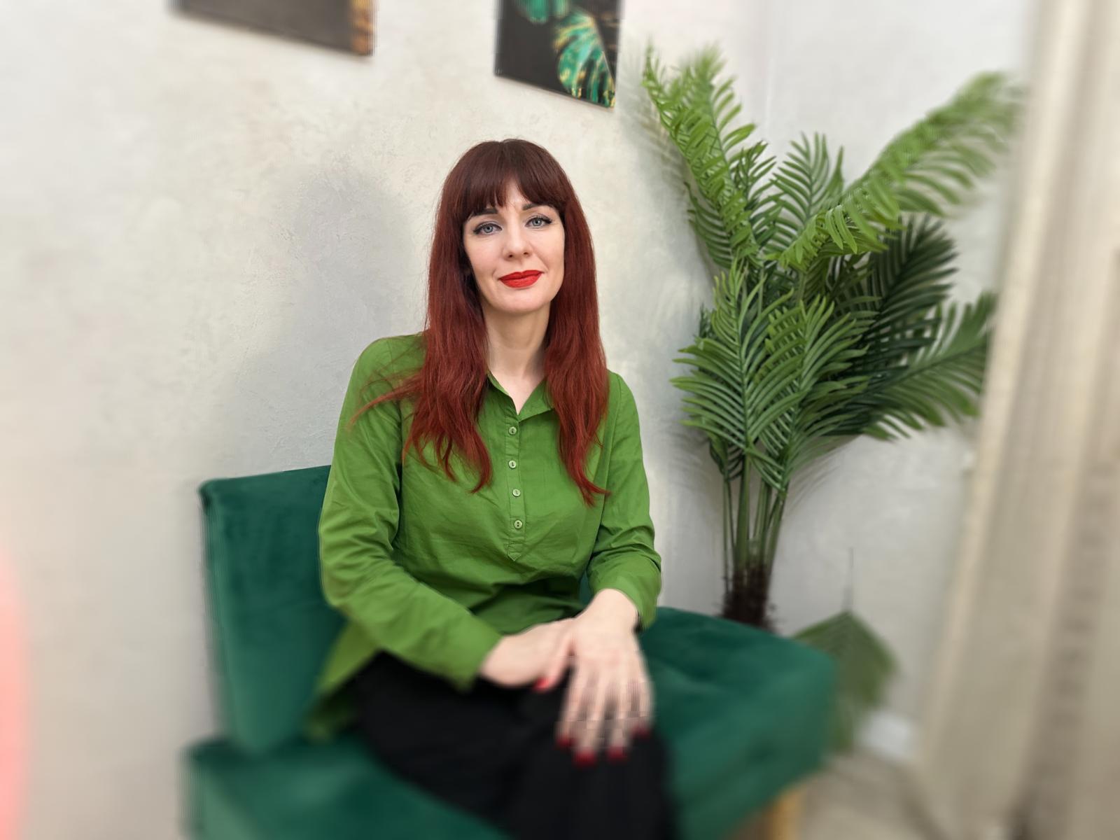 PureSeduce hat rote Haare, ein grünes Hemd, schwarze Hosen und roten Lippenstift, sitzt auf einem grünen Stuhl mit den Händen im Schoß, neben einer Topfpflanze und lächelt..
