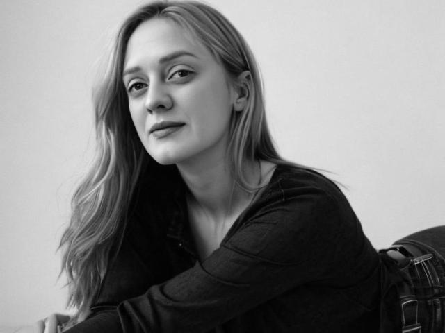 Black and white photo of KiraHarra with long, wavy hair, wearing a dark, long-sleeve shirt, and a plaid backpack, sitting with a slight, confident smile.