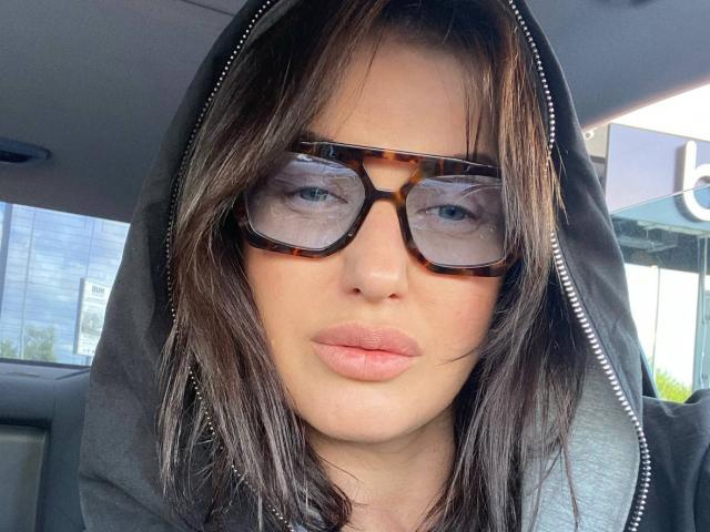 MeganSylvia with tortoiseshell glasses, wearing a grey hoodie, has light pink pouty lips, and brown straight hair, looking serious inside a car.
