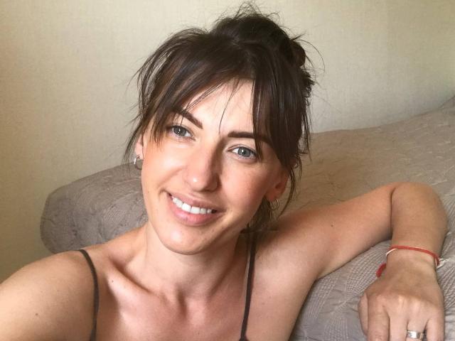 MeganSylvia with messy brown hair, green eyes, and a smile, wearing a black spaghetti strap top, showing her collarbone and a red bracelet on her right wrist.