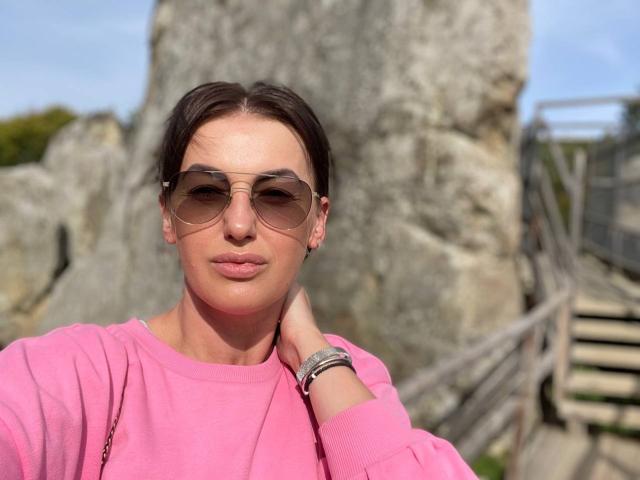 MeganSylvia with short brown hair, wearing aviator sunglasses, pink sweater, and bracelets, stands in front of rocky background with wooden stairs, slightly pouting lips.