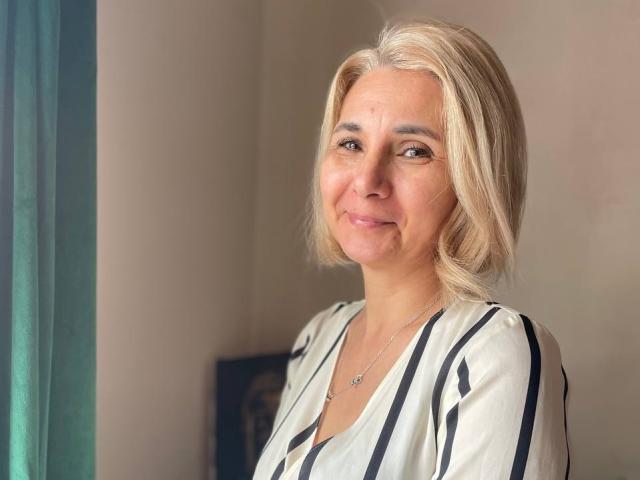 MiaRossy, com cabelo loiro, sorrindo suavemente, vestindo uma blusa listrada branca e preta e um colar pequeno, está em pé perto de uma cortina verde. Ela é uma mulher de meia-idade, muito charmosa..