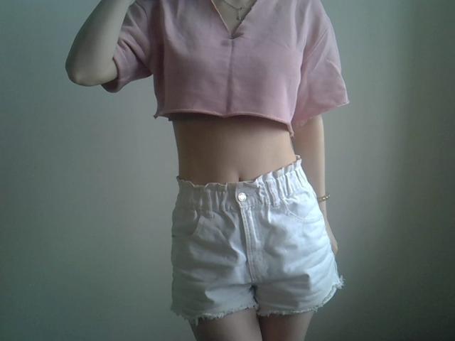 DitaVira wears a pink, short-sleeved crop top with a V-neck and frayed edges, exposing her midriff. She has white, high-waisted denim shorts with a button and frayed hem. Her left arm is raised, partially obscuring her face. The image focuses on her torso and lower body. She wears a thin bracelet on her right wrist. The light is soft, highlighting her skin and the textures of the clothing.
