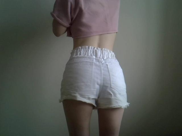 A photograph of DitaVira from behind. She wears a pink, cropped, short-sleeve shirt and high-waisted, white, frayed denim shorts that sit just above her knees. Her skin is light, and her back is slightly curved. The shorts have a loose, elastic waistband that adds a slight texture to the otherwise smooth fabric. The image focuses on her lower back and shorts, with her arms out of frame. The background is a plain, grayish wall.