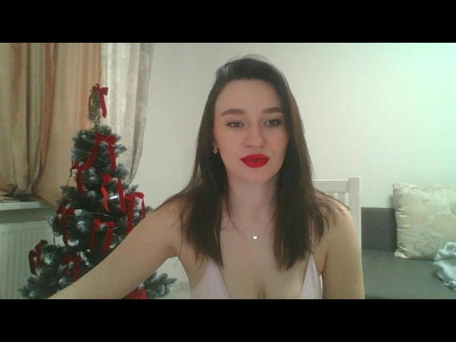 RozaSunn with red lipstick, medium tits, in a pink dress, standing next to a small Christmas tree with red ribbons, in a room with beige curtains.