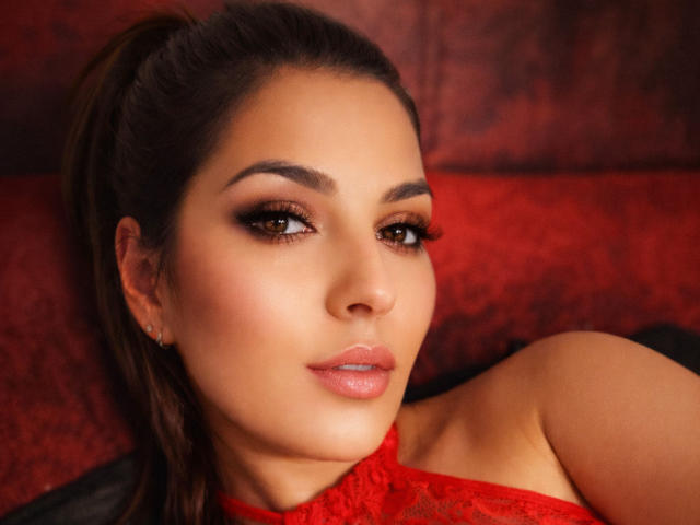 JolieBombonFontaine has a sexy face with brown eyes, full lips, and dark brown hair in a ponytail, wearing a red lace top, looking seductive with a slight smile.