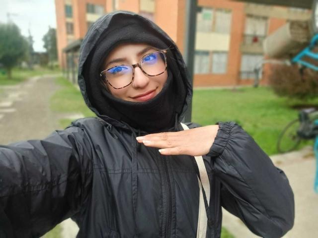 Kaiix with blue-tinted glasses, black hood, and puffy black jacket, smiling, hand on chest, black face mask, medium tits, slim, outside, overcast, green grass, bike in background.