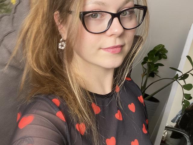KarKutie has light brown hair, black glasses, pink lipstick, and a black sheer top with red heart patterns, showing her face and upper chest. She's wearing a flower earring.