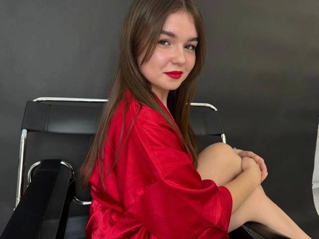 SofyX-hot with long brown hair, red lipstick, and red satin robe, sitting on black leather chair, knees up, smiling, showing her bare ass, in a studio shot.