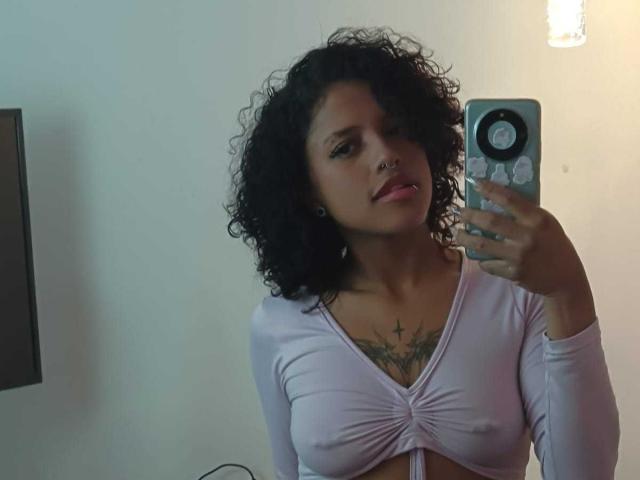 LunaKeller with curly black hair, nose ring, and tattooed chest, wearing a light purple, tied-up top, taking a mirror selfie, showing her small, perky tits.